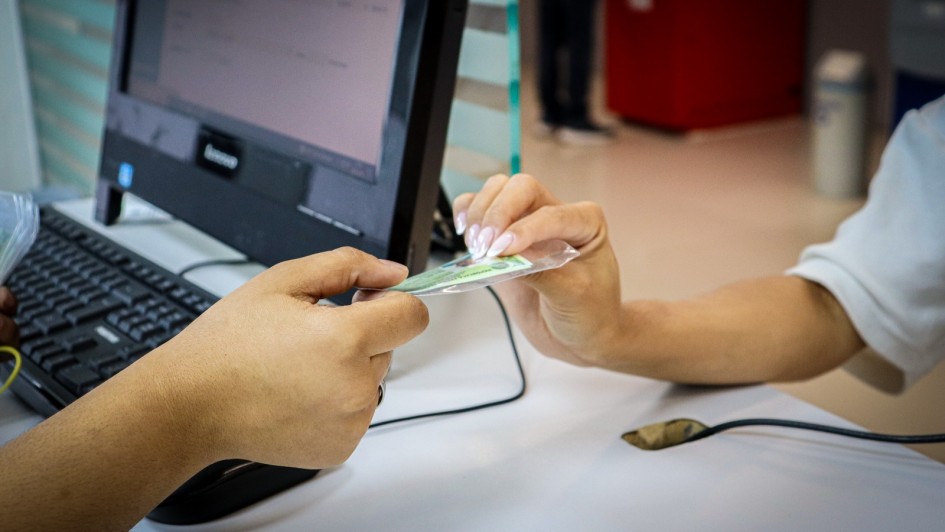 Duas mãos trocam um cartão plastificado diante de um computador ligado, com teclado sobre a mesa — uma cena comum de atendimento ou registro administrativo.