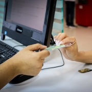 Duas mãos trocam um cartão plastificado diante de um computador ligado, com teclado sobre a mesa — uma cena comum de atendimento ou registro administrativo.