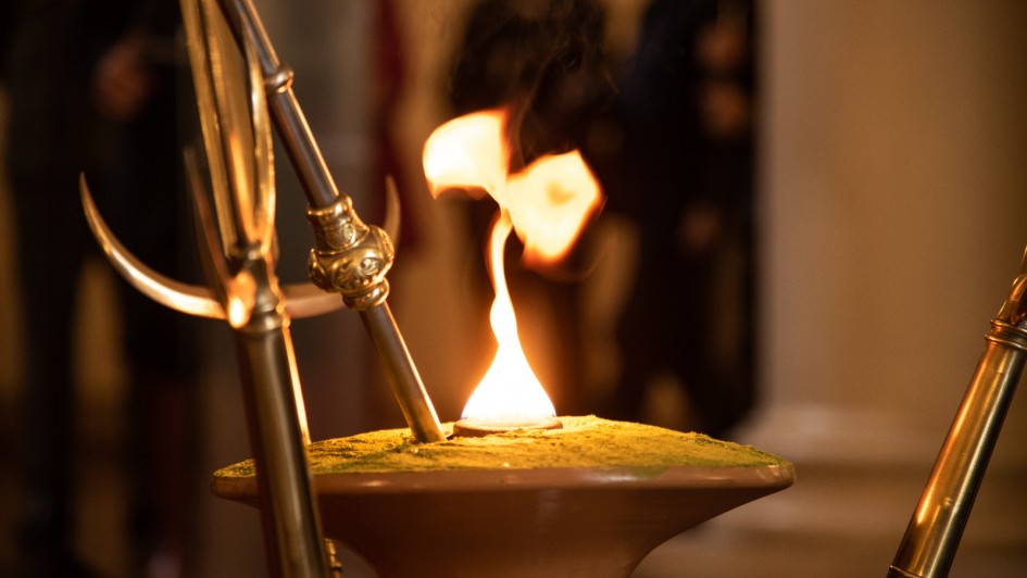 A imagem mostra uma chama acesa em uma espécie de recipiente cerâmico de boca larga, com o topo coberto por um pó de cor amarela-esverdeada. A chama está cercada por lanças ou instrumentos de metal com detalhes dourados, típicos da cultura gaúcha, simbolizando solenidade e tradição. Ao fundo, fora de foco, é possível ver pessoas observando a cerimônia em um ambiente interno, provavelmente um evento formal relacionado à cultura tradicionalista do Rio Grande do Sul. A iluminação quente destaca a chama como o elemento central da cena.