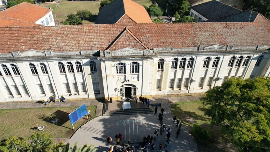 Foto aérea de um prédio histórico verticalizado, de cor clara e telhas alaranjadas. Há muitas janelas no edifício, e uma porta central que dá para uma escadaria e um pequeno pátio. Ao redor do pátio, há árvores. 