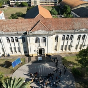 Foto aérea de um prédio histórico verticalizado, de cor clara e telhas alaranjadas. Há muitas janelas no edifício, e uma porta central que dá para uma escadaria e um pequeno pátio. Ao redor do pátio, há árvores. 