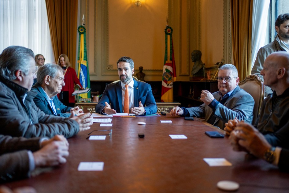 Reunião formal em uma sala elegante: várias pessoas sentadas ao redor de uma mesa grande, com papéis e eletrônicos em discussão. Bandeira do Brasil ao fundo, decoração clássica e ambiente sério indicam encontro oficial.