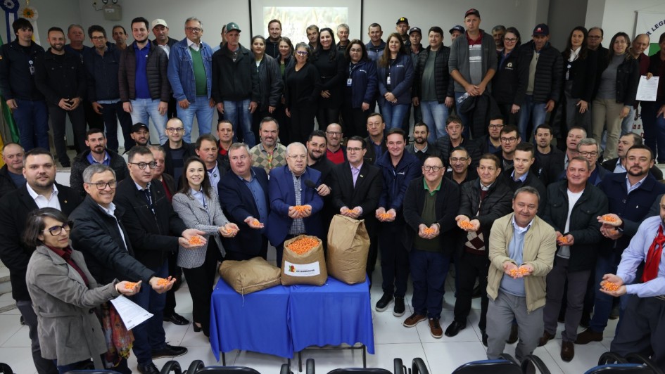 Imagem em ambiente interno. Há muitas pessoas em pé, reunidas lado a lado, posando para a foto. Na primeira fila, o titular da SDR e outros homens e mulheres seguram sementes de milho nas mãos enquanto sorriem e olham para a câmera. Ao centro há uma mesa com três sacos de sementes. 