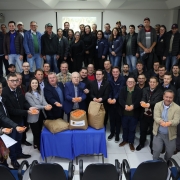Imagem em ambiente interno. Há muitas pessoas em pé, reunidas lado a lado, posando para a foto. Na primeira fila, o titular da SDR e outros homens e mulheres seguram sementes de milho nas mãos enquanto sorriem e olham para a câmera. Ao centro há uma mesa com três sacos de sementes. 