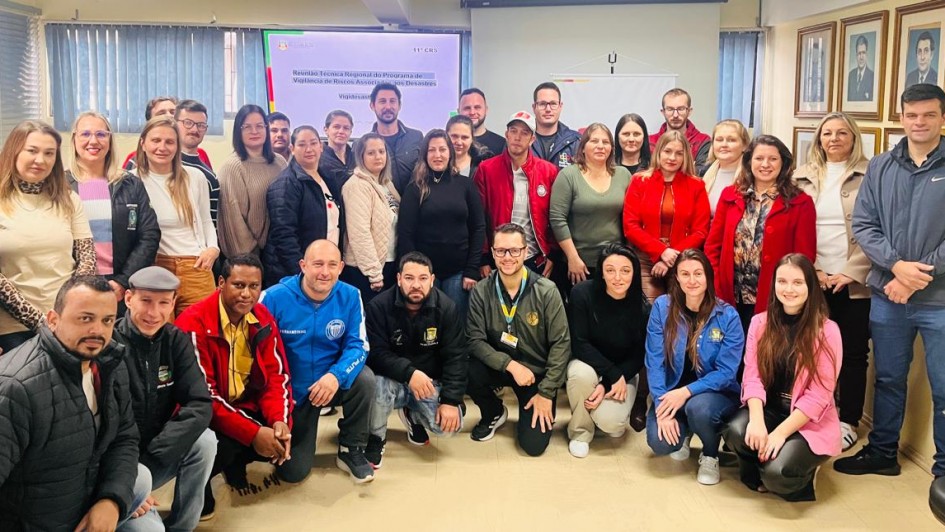 A foto mostra um grupo diversificado de aproximadamente trinta e cinco pessoas, entre homens e mulheres de várias idades, posando para uma foto em um ambiente interno que parece ser uma sala de reuniões ou auditório. A maioria das pessoas está em pé na parte de trás, enquanto alguns estão agachados na frente. Todos sorriem, olhando para a câmera. Ao fundo, uma tela ou projetor exibe um título de apresentação, e há banners e quadros decorando as paredes claras, sugerindo um evento de capacitação ou reunião profissional.
