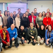 A foto mostra um grupo diversificado de aproximadamente trinta e cinco pessoas, entre homens e mulheres de várias idades, posando para uma foto em um ambiente interno que parece ser uma sala de reuniões ou auditório. A maioria das pessoas está em pé na parte de trás, enquanto alguns estão agachados na frente. Todos sorriem, olhando para a câmera. Ao fundo, uma tela ou projetor exibe um título de apresentação, e há banners e quadros decorando as paredes claras, sugerindo um evento de capacitação ou reunião profissional.