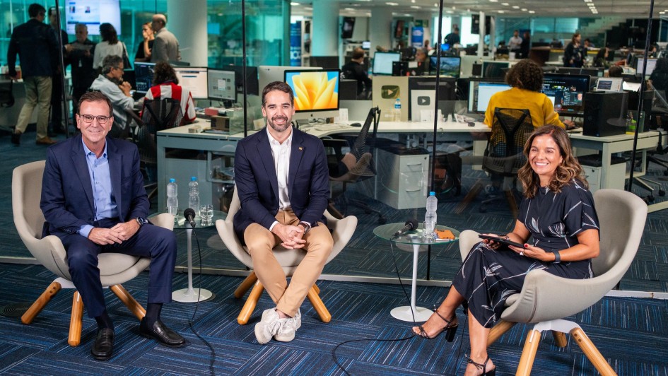 Imagem em um ambiente interno com mesas e computadores. O governador Eduardo Leite está posicionado em destaque, ao lado de um homem e uma mulher. Os três estão sentados em poltronas e sorriem para a câmera. 