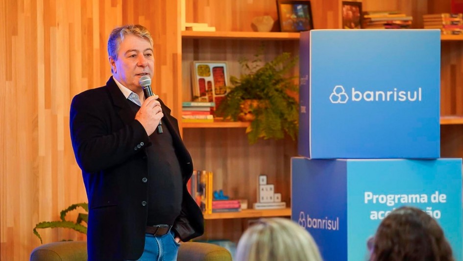 Homem de cabelos grisalhos, vestindo blazer preto e camisa azul clara, está em pé segurando um microfone enquanto fala a um público. Ele está posicionado à esquerda da imagem. À direita, há dois cubos grandes com o logotipo do Banrisul e os dizeres "Programa de aceleração". Ao fundo, prateleiras de madeira com livros, plantas e objetos decorativos compõem o ambiente, que tem aparência moderna e acolhedora. Algumas pessoas aparecem de costas na parte inferior da imagem, compondo a plateia.