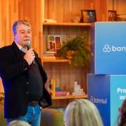 Homem de cabelos grisalhos, vestindo blazer preto e camisa azul clara, está em pé segurando um microfone enquanto fala a um público. Ele está posicionado à esquerda da imagem. À direita, há dois cubos grandes com o logotipo do Banrisul e os dizeres "Programa de aceleração". Ao fundo, prateleiras de madeira com livros, plantas e objetos decorativos compõem o ambiente, que tem aparência moderna e acolhedora. Algumas pessoas aparecem de costas na parte inferior da imagem, compondo a plateia.