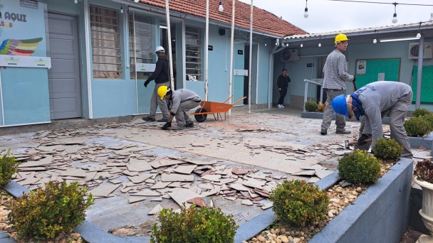A foto mostra um pátio externo em reforma na EEEM Catarina Bridi, onde trabalhadores com capacetes amarelos estão removendo detritos de construção. O chão está coberto por pedaços de telhas quebradas e entulho, com um carrinho de mão laranja vazio no centro. Os trabalhadores, vestidos com uniformes cinzas, agacham-se para recolher o material. Ao fundo, uma parte do prédio da escola, com paredes claras e janelas, é visível, junto com arbustos e um céu claro. A cena transmite o trabalho em andamento para melhorias na infraestrutura.