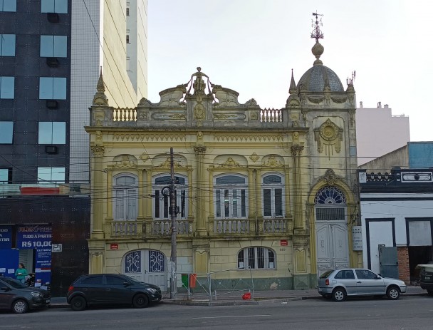 A foto mostra a fachada imponente e antiga da EEEM Nossa Senhora de Lourdes, um edifício amarelo ornamentado com detalhes em relevo e balaustradas, típico de arquitetura histórica. Possui várias janelas com molduras brancas e uma porta de entrada em arco com uma cúpula elaborada e uma cata-vento no topo. Carros estão estacionados na rua em frente ao prédio, e edifícios modernos mais altos são visíveis ao fundo, criando um contraste entre o antigo e o novo. A iluminação é clara, e o céu, azul claro, realça a arquitetura.