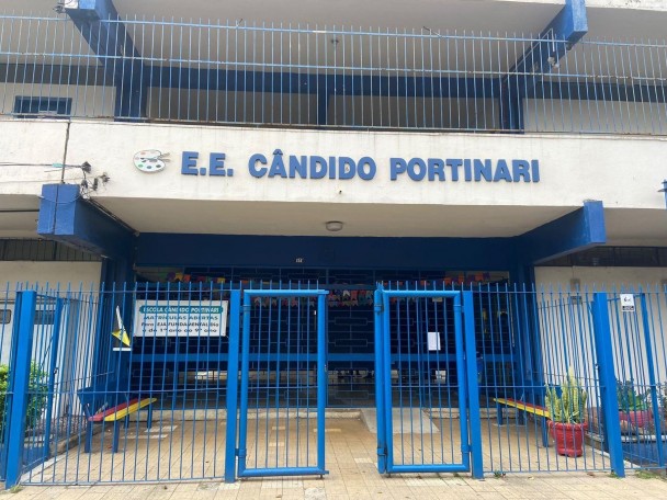 A foto mostra a fachada principal da EEEF Cândido Portinari, um prédio de dois andares com paredes brancas e detalhes em azul. Uma grande grade azul com portões centrais fecha a entrada da escola, onde há um aviso sobre matrículas abertas. O nome da escola está escrito em letras grandes e azuis na parte superior do prédio, acompanhado de um pequeno logotipo colorido. Vasos de plantas e um banco simples são visíveis atrás da grade. A iluminação é uniforme, e o céu parece claro, sugerindo um dia tranquilo.