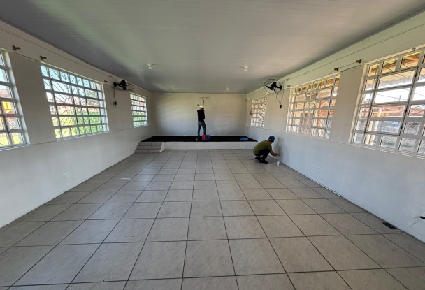 A foto mostra um salão amplo e vazio dentro da EEEF Coronel Juvêncio Lemos, com paredes recém-pintadas de branco e um piso de lajotas bege. Há várias janelas altas com grades, permitindo a entrada de luz natural. Dois trabalhadores estão visíveis na sala: um homem de camiseta escura está de costas no fundo do salão, e outro, com capacete azul e uniforme, agacha-se perto da parede direita, pintando a parte inferior. O espaço parece estar passando por reformas ou preparativos para uso, com ventiladores de parede visíveis.