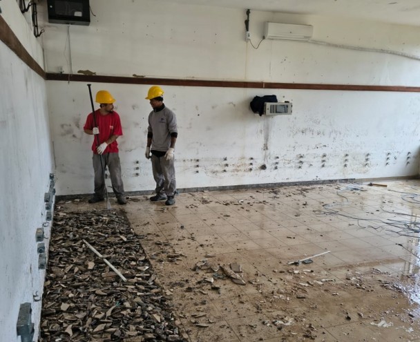 A foto mostra o interior de uma sala em reforma na EEEF Jovelino Theodoro, com o piso em processo de remoção. Dois trabalhadores, ambos usando capacetes amarelos, estão em pé no centro da sala. Um deles, à esquerda, com camiseta vermelha e luvas, segura uma ferramenta longa e está de frente para um monte de detritos no chão. O outro, à direita, de camiseta cinza, observa. O piso antigo está quebrado, e cabos elétricos expostos saem da parede, indicando uma obra de grande porte.