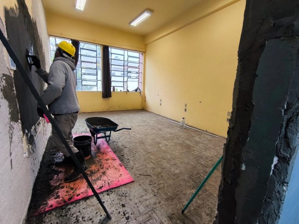 A foto mostra um trabalhador com capacete amarelo aplicando massa em uma parede interna, em uma sala em reforma na EEEF Santos Dumont. O homem, visto de costas, veste uma jaqueta cinza clara e calça escura, com luvas rosas. Ferramentas e um balde de argamassa estão próximos. O chão da sala é de tijolos e está coberto por detritos e um plástico rosa. As paredes são claras, e uma janela grande permite a entrada de luz natural. Um carrinho de mão e materiais de construção são visíveis, indicando o andamento da obra.