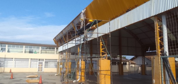 A foto mostra uma grande estrutura de telhado metálico corrugado em tom amarelado, provavelmente de um ginásio ou galpão, em processo de reforma no IE Barão de Tramandaí. Andaimes de metal com telas de proteção envolvem parte da estrutura, e um trabalhador com capacete amarelo é visível sobre eles, realizando reparos. Ao fundo, um prédio escolar de um andar com paredes claras e muitas janelas com grades é visível, junto com um pátio de cimento. O céu é azul com poucas nuvens, indicando um dia ensolarado, e cones de sinalização laranja estão no chão.