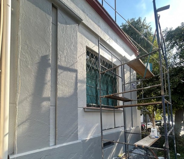 A foto mostra a lateral de um edifício em processo de reforma, provavelmente o CE Inácio Montanha. A parede, de cor clara, parece estar recém-pintada ou com aplicação de textura, exibindo algumas manchas mais escuras de umidade ou sujeira na parte inferior. Uma janela com grades de ferro está parcialmente visível, protegida por andaimes de metal. Uma tábua de madeira está apoiada nos andaimes, e um balde com materiais de construção está no chão. Árvores e o céu azul com algumas nuvens são visíveis ao fundo, indicando um dia ensolarado.
