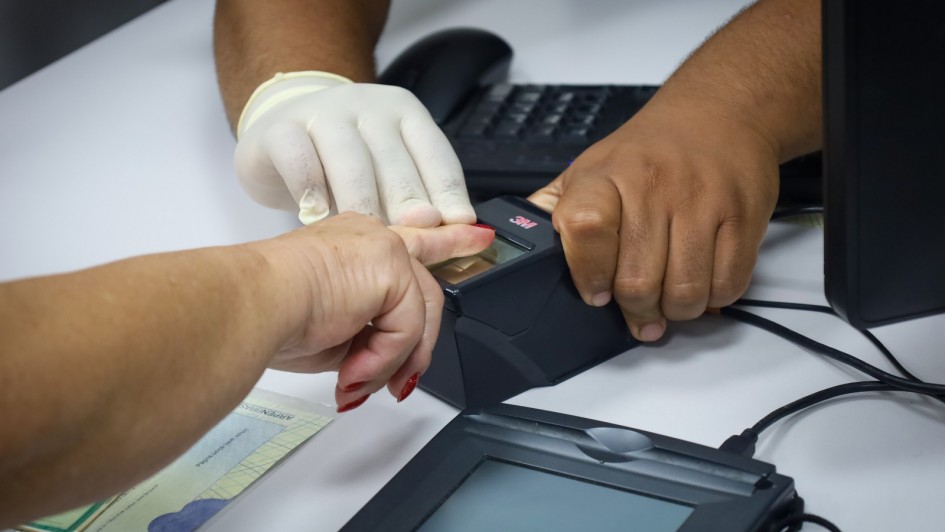 Uma pessoa está tendo sua impressão digital coletada por um leitor biométrico, com o auxílio de um atendente. A mão da pessoa está sendo pressionada sobre o sensor digital por um atendente que usa uma luva descartável em uma das mãos. Sobre a mesa branca, ao lado do leitor biométrico, há um documento de identidade parcialmente visível e um teclado de computador. A cena parece ocorrer em um ambiente de atendimento público, como um posto de identificação civil ou cartório.