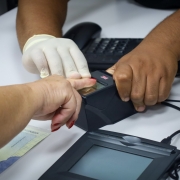 Uma pessoa está tendo sua impressão digital coletada por um leitor biométrico, com o auxílio de um atendente. A mão da pessoa está sendo pressionada sobre o sensor digital por um atendente que usa uma luva descartável em uma das mãos. Sobre a mesa branca, ao lado do leitor biométrico, há um documento de identidade parcialmente visível e um teclado de computador. A cena parece ocorrer em um ambiente de atendimento público, como um posto de identificação civil ou cartório.