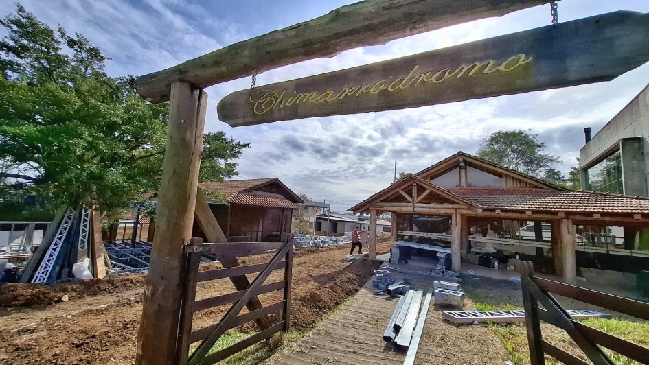 A foto mostra uma área do Parque Assis Brasil em reforma, com foco em uma estrutura rústica de madeira que serve como um portal de entrada, exibindo uma placa com a palavra "Chimarródromo" em caligrafia elaborada. O chão ao redor do portal está em terra batida, com algumas tábuas no caminho. Ao fundo, diversas edificações com telhados de madeira estão em construção ou reforma, com materiais de obra espalhados no chão, incluindo estruturas metálicas e tubos. O céu está parcialmente nublado, e árvores completam a paisagem.
