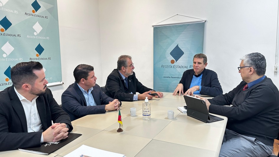 Cinco homens estão sentados ao redor de uma mesa retangular em uma sala de reuniões da Receita Estadual do RS. Eles conversam de forma atenta, com papéis, canetas, notebook e uma pequena bandeira do Rio Grande do Sul sobre a mesa. Ao fundo, dois banners com o logotipo da Receita Estadual decoram o ambiente.