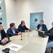 Cinco homens estão sentados ao redor de uma mesa retangular em uma sala de reuniões da Receita Estadual do RS. Eles conversam de forma atenta, com papéis, canetas, notebook e uma pequena bandeira do Rio Grande do Sul sobre a mesa. Ao fundo, dois banners com o logotipo da Receita Estadual decoram o ambiente.