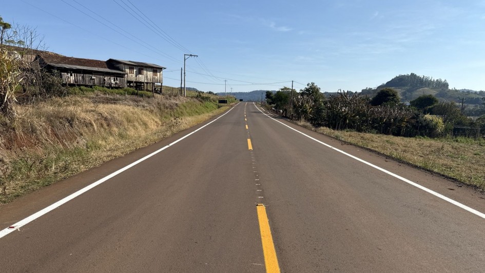 A imagem mostra uma estrada asfaltada de mão dupla em área rural, com faixa central amarela contínua e laterais brancas. O céu está limpo e azul. À esquerda da estrada, há uma casa antiga de madeira em estado de desgaste, localizada em um pequeno barranco com vegetação seca. À direita, há vegetação mais densa, incluindo árvores e plantas, com colinas ao fundo. A estrada segue em linha reta em direção ao horizonte, com postes de energia ao longo da via.