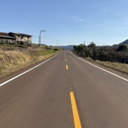 A imagem mostra uma estrada asfaltada de mão dupla em área rural, com faixa central amarela contínua e laterais brancas. O céu está limpo e azul. À esquerda da estrada, há uma casa antiga de madeira em estado de desgaste, localizada em um pequeno barranco com vegetação seca. À direita, há vegetação mais densa, incluindo árvores e plantas, com colinas ao fundo. A estrada segue em linha reta em direção ao horizonte, com postes de energia ao longo da via.