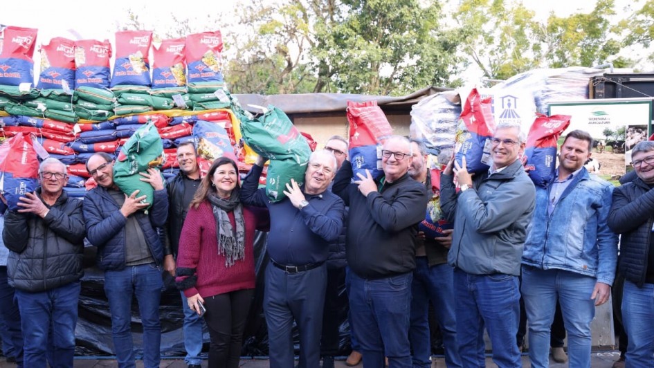 Imagem ao ar livre. Diversas pessoas estão sorrindo para a foto, em pé, segurando sacas de sementes nas cores verde, vermelha e azul. 