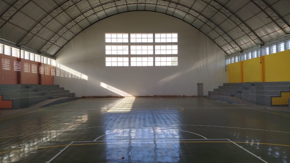 A foto mostra o interior de um ginásio poliesportivo, que é o Ginásio do IE Cristóvão de Mendoza. O espaço é amplo, com um teto abobadado de estrutura metálica aparente e redes de proteção nas laterais. No centro, a quadra de esportes tem piso em tons de verde e faixas amarelas e brancas, delimitando as áreas de jogo. Há algumas pequenas manchas de sujeira no chão. As arquibancadas, de cor cinza e laranja, estão vazias e elevadas nas laterais. No fundo, uma grande janela retangular no alto da parede permite a entrada de luz natural, que forma feixes e reflexos no chão, destacando a poeira no ar. A iluminação é mista, com a luz natural e algumas lâmpadas no teto.