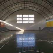 A foto mostra o interior de um ginásio poliesportivo, que é o Ginásio do IE Cristóvão de Mendoza. O espaço é amplo, com um teto abobadado de estrutura metálica aparente e redes de proteção nas laterais. No centro, a quadra de esportes tem piso em tons de verde e faixas amarelas e brancas, delimitando as áreas de jogo. Há algumas pequenas manchas de sujeira no chão. As arquibancadas, de cor cinza e laranja, estão vazias e elevadas nas laterais. No fundo, uma grande janela retangular no alto da parede permite a entrada de luz natural, que forma feixes e reflexos no chão, destacando a poeira no ar. A iluminação é mista, com a luz natural e algumas lâmpadas no teto.