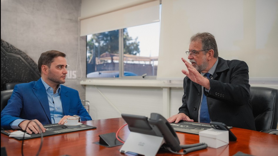 Em uma sala de reuniões, dois homens estão sentados à mesa. Ganbriel, de terno azul, ouve atentamente. O CEO da Taurus, de terno escuro e gravata azul, gesticula com a mão levantada enquanto fala. Há telefones e papéis sobre a mesa.