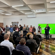 A foto mostra o Secretário da Agricultura, Edivilson Brum, um homem de pele clara e cabelo escuro, em pé no centro da imagem. Ele veste uma jaqueta escura e está segurando um microfone, falando para uma plateia. Ao seu lado direito, um telão verde exibe alguns logotipos. À esquerda do secretário, há um grupo de cerca de dez pessoas em pé, incluindo homens e mulheres, olhando para a plateia. Em primeiro plano, uma plateia de aproximadamente trinta pessoas, a maioria homens de meia-idade, está sentada em cadeiras escuras, com muitos de costas para a câmera. O ambiente é uma sala ampla com teto branco e iluminação fluorescente, e no fundo, uma porta de madeira e quadros na parede.