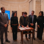 A foto mostra um grupo de seis pessoas em pé ao redor de uma pequena mesa de madeira, em um ambiente interno formal. No centro, o governador em exercício, Gabriel Souza, com barba, vestido com um terno marrom e camisa escura, está assinando um documento. Ele está inclinado sobre a mesa, com a caneta na mão, e todos os demais o observam com atenção. Ao lado esquerdo de Gabriel, uma mulher de cabelo escuro e camisa preta está ao seu lado. O resto do grupo é composto por quatro homens de terno. O ambiente tem paredes claras e uma janela alta com cortinas. Ao fundo, um telão mostra uma apresentação e, à direita, há uma estátua e uma coluna. O chão é de madeira.
