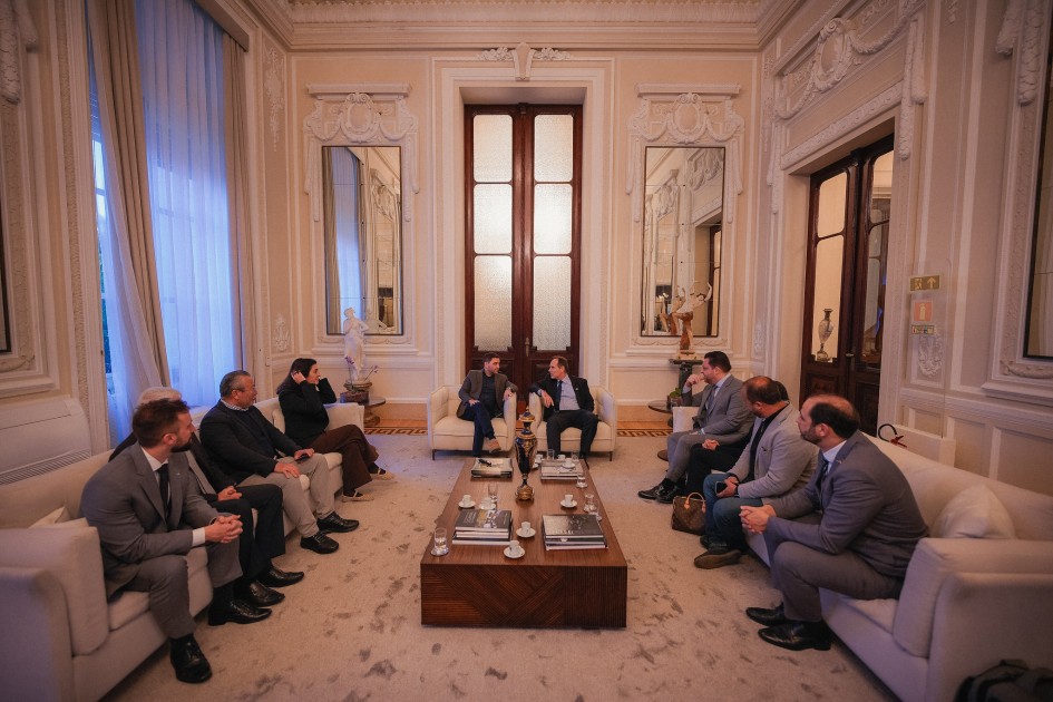 A foto mostra um grupo de oito homens sentados em sofás e poltronas brancas, dispostos em um grande círculo em uma sala de estar formal. O ambiente tem teto alto e detalhes arquitetônicos em gesso, com grandes espelhos e janelas com cortinas longas. Os homens estão em trajes sociais, conversando e interagindo uns com os outros. A iluminação é clara e o ambiente parece luxuoso.