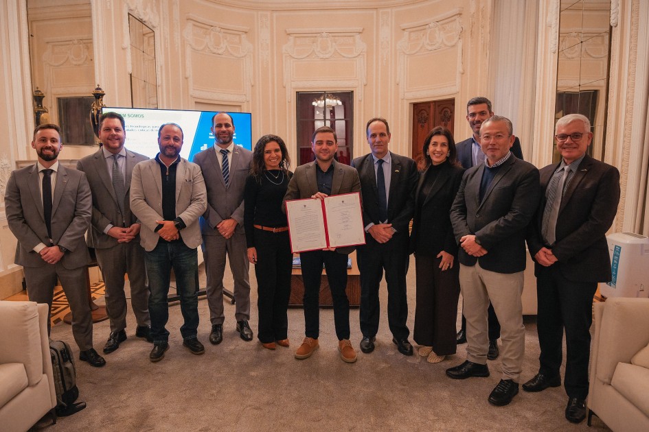 A foto mostra um grupo de dez pessoas, a maioria homens e três mulheres, em pé e posando para uma foto em um salão com arquitetura clássica. O grupo está alinhado em frente a uma tela onde se vê um telão, e um dos homens, no centro, o governador em exercício, Gabriel Souza, segura um documento com bordas vermelhas. Ele veste terno escuro. O ambiente tem paredes claras, um teto com detalhes em gesso e grandes janelas com cortinas. A iluminação é uniforme, vinda do alto.