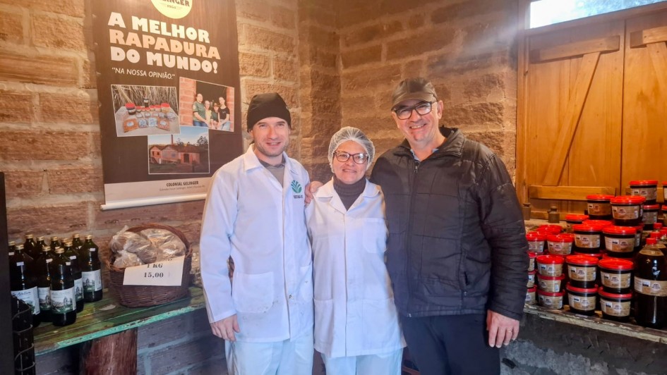 Três pessoas estão sorrindo e posando para a foto dentro de um ambiente rústico, com paredes de tijolos aparentes e uma janela de madeira ao fundo. À direita, um homem veste jaqueta preta e boné escuro. Ao centro, uma mulher usa jaleco branco, touca e óculos. À esquerda, um jovem também com jaleco branco e touca preta. Atrás deles, há um cartaz com os dizeres: “A MELHOR RAPADURA DO MUNDO! – NA NOSSA OPINIÃO”, com fotos da produção e da família. À esquerda da imagem, há garrafas e uma cesta com produtos etiquetada com o preço “1KG – 15,00”. À direita, potes empilhados com tampas vermelhas.