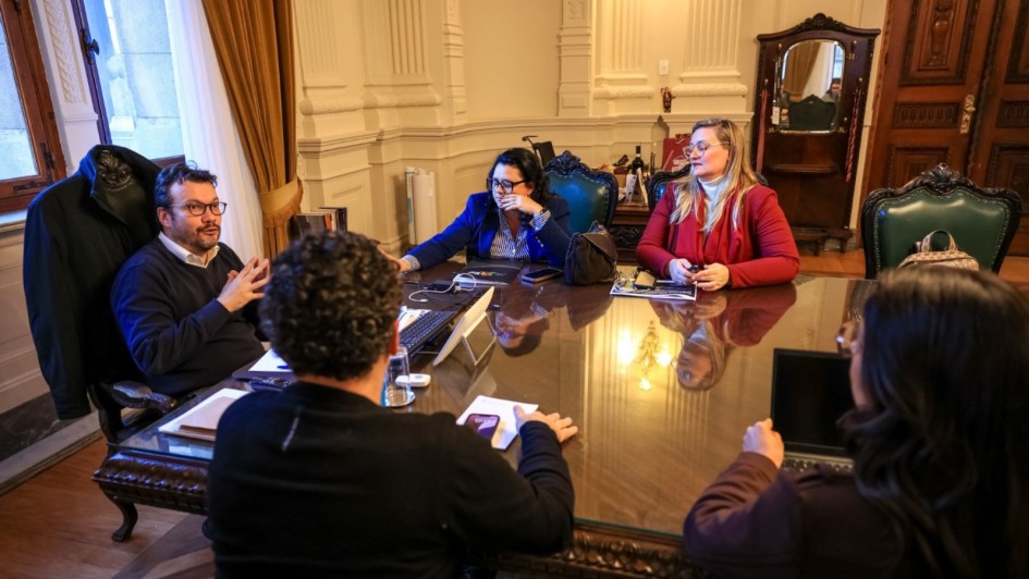 A foto mostra quatro pessoas sentadas em uma mesa de reunião grande e retangular, com tampo de madeira, em uma sala com arquitetura clássica. A secretária do Meio Ambiente e Infraestrutura, Marjorie Kauffmann, está sentada à frente e à esquerda, vestindo um casaco azul e com o cotovelo apoiado na mesa, olhando para o lado. À sua frente, um homem de blusa escura e óculos, que é o Chefe da Casa Civil, Artur Lemos, está sentado na ponta da mesa. Do outro lado da mesa, um homem com barba e óculos, com um suéter azul, está sentado à esquerda e interagindo. Uma quarta mulher de blusa vermelha e cabelos loiros está sentada à direita. A mesa tem papéis, tablets, xícaras de café e notebooks. A sala tem teto com adornos, janelas com cortinas longas e paredes com molduras.