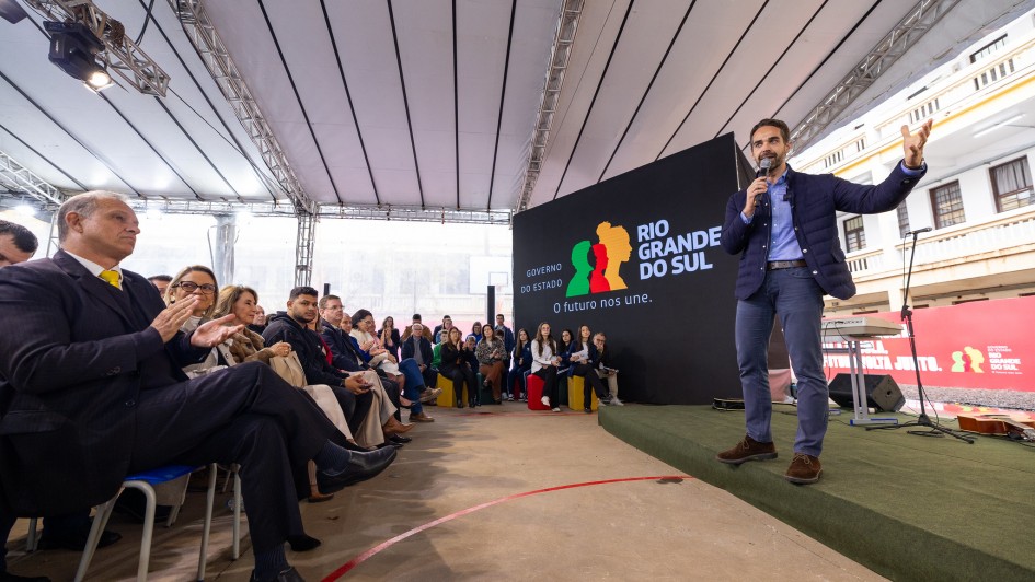 Fotografia de um evento realizado no pátio coberto do Colégio Estadual Presidente Castelo Branco, em Lajeado. O governador do Rio Grande do Sul, segurando um microfone, faz discurso diante da plateia. À esquerda, autoridades sentadas acompanham e aplaudem. O telão e a estrutura coberta continuam visíveis, assim como parte do prédio escolar ao fundo.