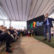 Fotografia de um evento realizado no pátio coberto do Colégio Estadual Presidente Castelo Branco, em Lajeado. O governador do Rio Grande do Sul, segurando um microfone, faz discurso diante da plateia. À esquerda, autoridades sentadas acompanham e aplaudem. O telão e a estrutura coberta continuam visíveis, assim como parte do prédio escolar ao fundo.
