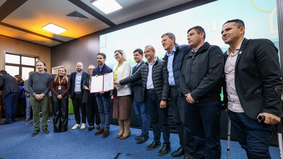 Pessoas reunidas em evento formal: grupo em pé, com roupas sociais ou casuais, ao redor de um grande livro ou documento aberto, destaque da cena. Ao fundo, tela luminosa com texto e painéis de madeira nas paredes, proporcionando ambiente moderno e bem iluminado.