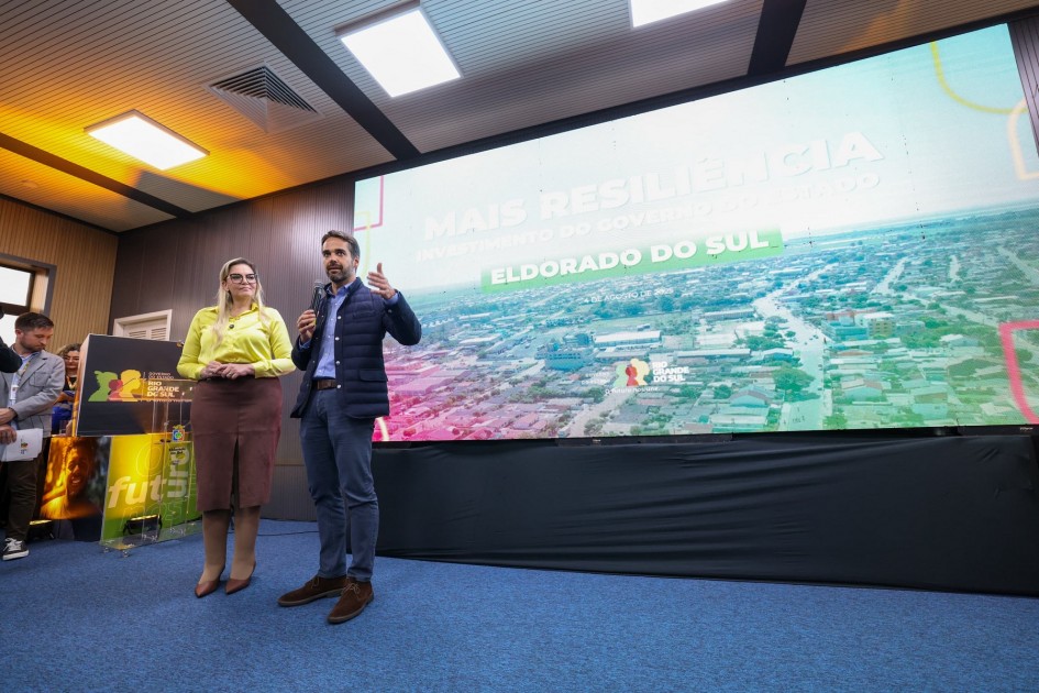 Evento público em destaque: duas pessoas no palco conversam ao microfone diante de uma tela grande que mostra uma imagem aérea da cidade. A apresentação é sobre investimentos do governo em Eldorado do Sul, com foco em resiliência e futuro. Ambiente com tapete azul, iluminação retangular no teto e sinalização oficial do Estado do Rio Grande do Sul.