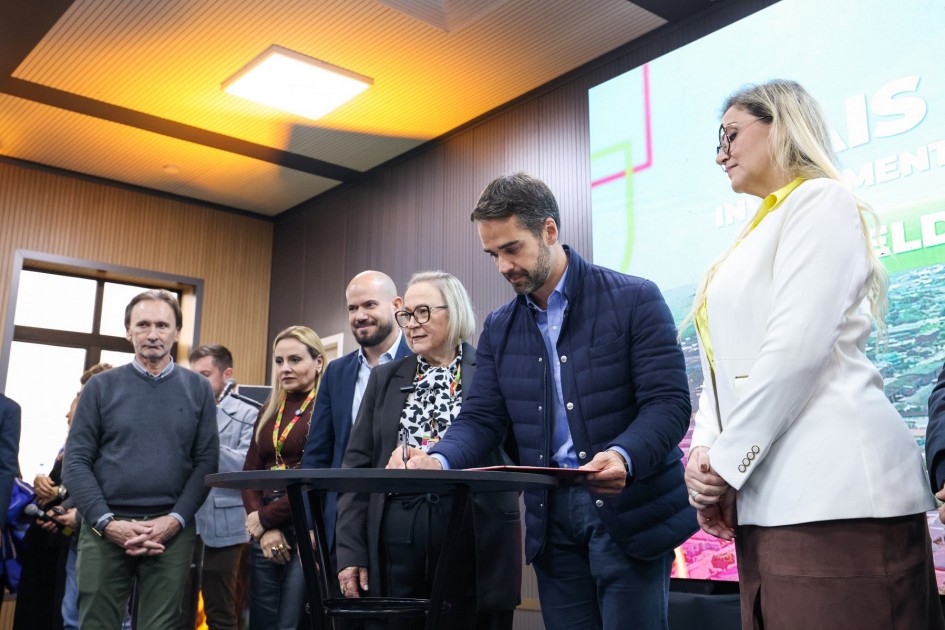 Momento de assinatura: pessoa em destaque escreve sobre uma mesa preta, cercada por observadores atentos. A sala tem paredes de madeira, iluminação clara no teto e uma grande tela ao fundo com palavras como “investimento” e “campo”, indicando um evento formal para oficializar parcerias ou decisões importantes.
