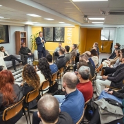 Esta é uma fotografia de um evento de treinamento ou apresentação em um ambiente de escritório ou sala de aula.

Em primeiro plano, a plateia é vista de costas, ocupando a maior parte da imagem. Homens e mulheres de diversas idades estão sentados em cadeiras de madeira, voltados para a frente da sala.

No centro da imagem, em pé, um homem de barba, usando uma jaqueta escura sobre uma camisa preta, está falando para o grupo. Ele parece estar segurando um pequeno controle remoto ou um microfone.

À esquerda, no fundo, uma mulher com cabelo escuro, vestindo uma camiseta preta, está de pé ao lado de uma tela de televisão grande que exibe um slide de apresentação com dados em colunas.

As paredes da sala são de cor cinza e amarela, com a palavra "LEARN" (aprender, em inglês) escrita em letras grandes e verticais em um pilar à direita. Há grandes janelas ao fundo da sala. O teto é branco e possui luminárias retangulares. A iluminação é clara e a imagem é nítida.