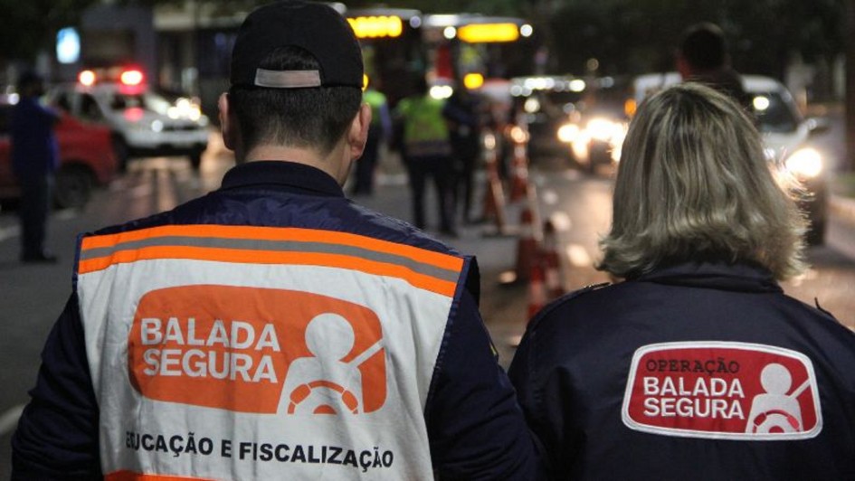 A foto mostra dois agentes de trânsito de costas em uma rua durante a noite. O agente à esquerda, um homem de boné preto, veste um colete branco com faixas laranjas refletivas, onde se lê "BALADA SEGURA" e "EDUCAÇÃO E FISCALIZAÇÃO" em letras pretas. A agente à direita, uma mulher de cabelos loiros, veste um casaco escuro com um distintivo "OPERAÇÃO BALADA SEGURA" nas costas. Ao fundo, carros com luzes de farol acesas e luzes vermelhas de emergência são visíveis, indicando que se trata de uma operação de trânsito em uma rua movimentada. A iluminação é proveniente dos veículos e das luzes da rua.