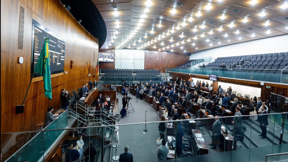 A foto mostra o plenário da Assembleia Legislativa do Rio Grande do Sul, um grande salão com paredes de madeira escura e um teto alto com iluminação moderna. No primeiro plano, uma escadaria leva a um andar superior, com um corrimão de vidro. No centro da sala, há um auditório com várias fileiras de cadeiras estofadas escuras, onde a plateia está sentada. Em uma área mais elevada, à esquerda, uma longa mesa retangular com cadeiras acomoda um grupo de pessoas, que parecem ser membros da mesa diretora ou de uma comissão. Ao fundo, uma grande tela de LED exibe informações. O ambiente está bem iluminado, e muitas pessoas, em sua maioria em trajes formais, estão presentes na sessão.