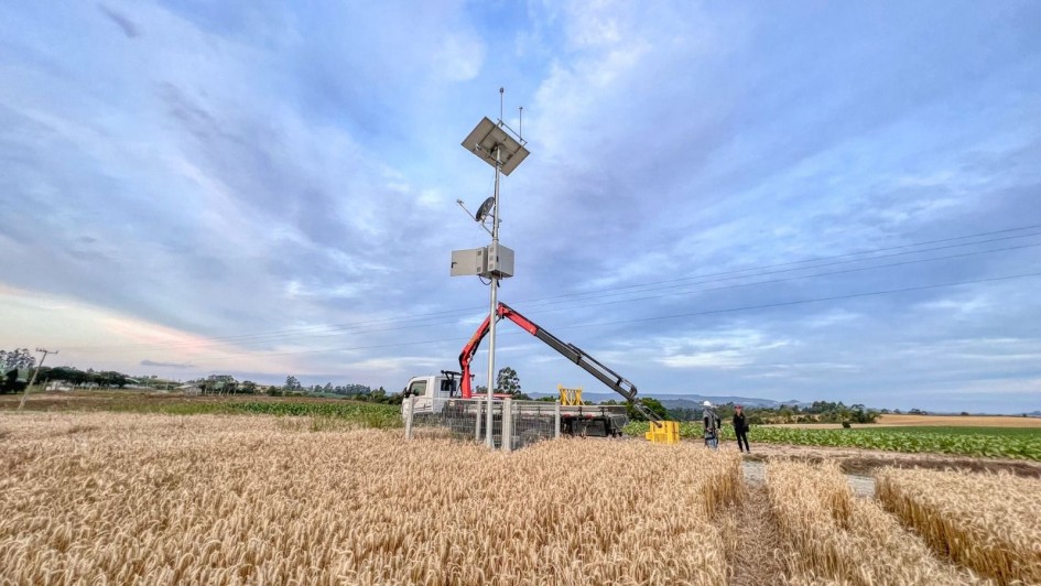 Foto ao ar livre, em um dia claro. Há uma estação meteorológica em cima de uma caminhão no meio de um campo. Por perto, há duas pessoas olhando para o equipamento.