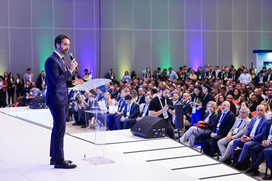 Foto em ambiente fechado. O governador Eduardo Leite está no palco, no canto esquerdo da imagem, falando ao microfone para uma plateia lotada.