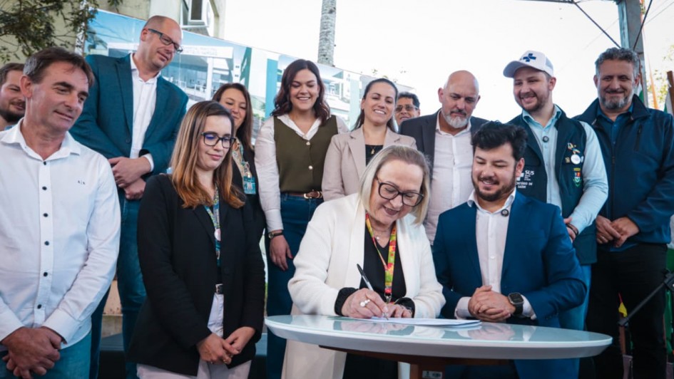A foto mostra o momento de assinatura do documento. A Secret&aacute;ria Arita Bergmann, com &oacute;culos e um casaco branco, est&aacute; em p&eacute;, sorrindo, enquanto assina um papel em uma mesa redonda e branca. Ao seu lado, um homem de terno azul-marinho e um homem de camisa branca e cal&ccedil;a jeans a observam. Outras pessoas, incluindo um homem com uma camisa polo preta e uma mulher com um blazer branco, est&atilde;o em volta, sorrindo e assistindo ao ato. O evento acontece em uma &aacute;rea externa, sob uma cobertura, com a presen&ccedil;a de outras pessoas ao fundo.