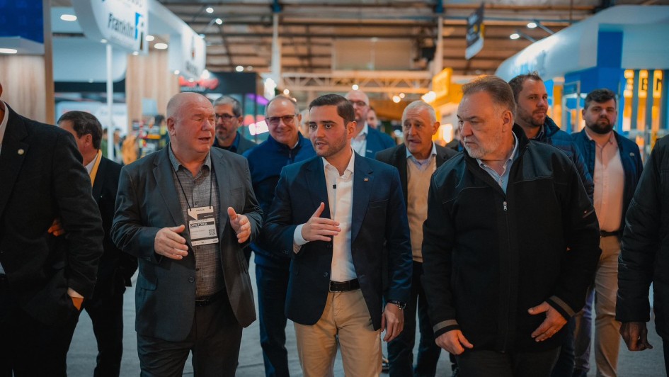 Um grupo de homens caminha por um corredor coberto de uma feira ou evento. No centro da imagem, três deles estão em destaque, conversando entre si. O homem do meio veste blazer azul-marinho, camisa branca e calça clara; ele parece liderar a comitiva. À esquerda, outro homem calvo com terno cinza e crachá conversa com ele, gesticulando com as mãos. À direita, um terceiro homem com barba grisalha veste jaqueta escura e observa a conversa. Ao fundo, outras pessoas acompanham o grupo e há estandes iluminados, com logotipos visíveis e iluminação artificial no teto, sugerindo um ambiente profissional e organizado, como uma feira de negócios ou exposição industrial.